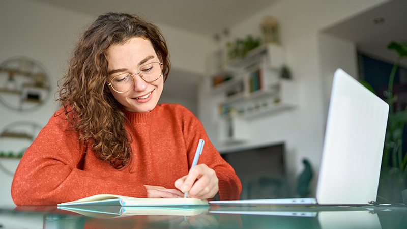 Mujer joven, escribiendo en un papel, con una computadora  en el escritorio